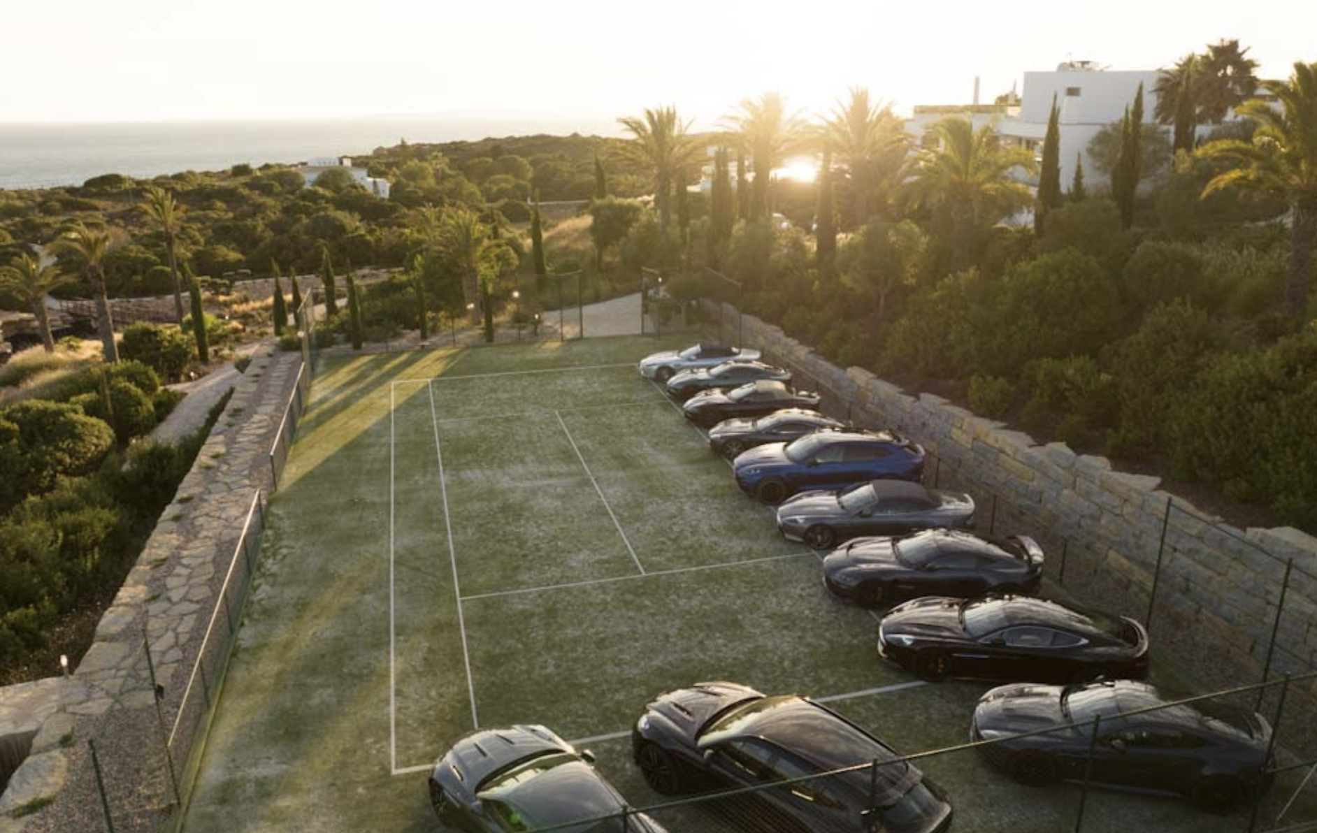 Grass tennis court at sunset with luxury sports cars lined along the stone wall